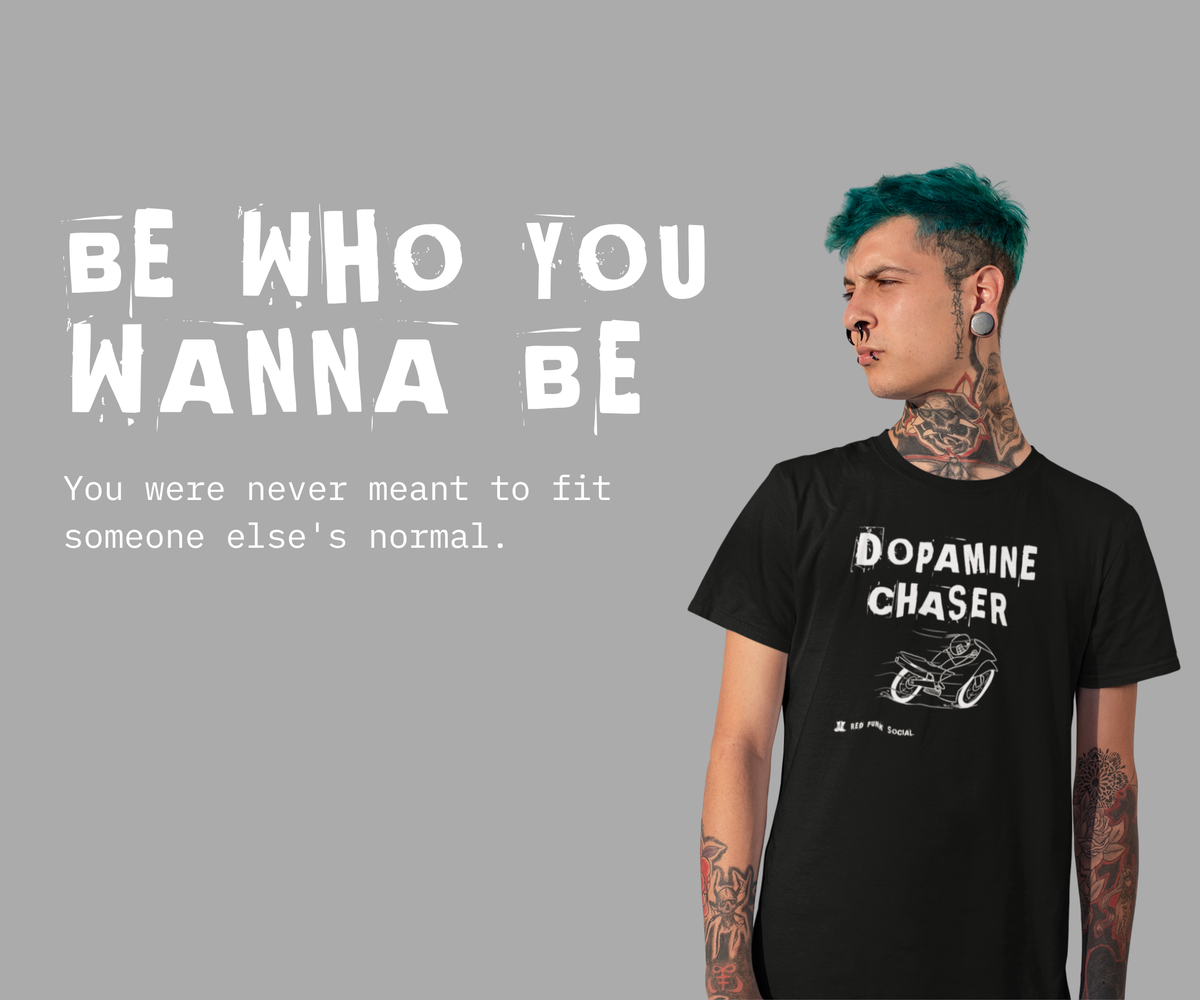 A man with teal green/blue hair stands in front of a grey background wearing a punk black T-shirt with white punk font written with the words: DOMPAMINE CHASER. There is a white speeding motorbike underneath the font and the Red Punk Social brand logo on the tee, in the bottom left corner. Left of the man are the words in large punk font “BE WHO YOU WANNA BE” beneath this in small type-font, “You were never meant to fit someone else’s normal.”
